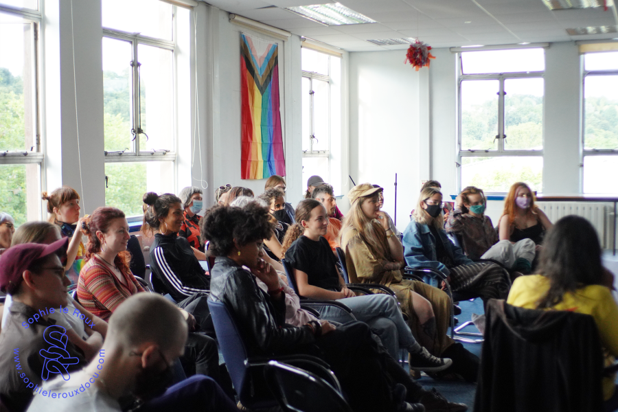 Lots of people sat in the blue Room listening, a pride flag hangs on the wall in the background