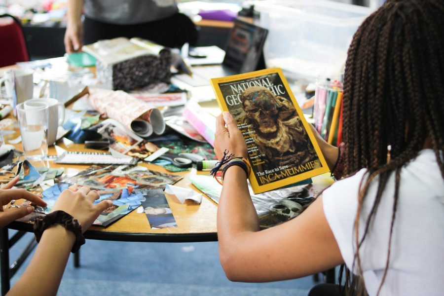 Two people's arms leaning over a table, holding onto magazines with images on the table, at a collage workshop