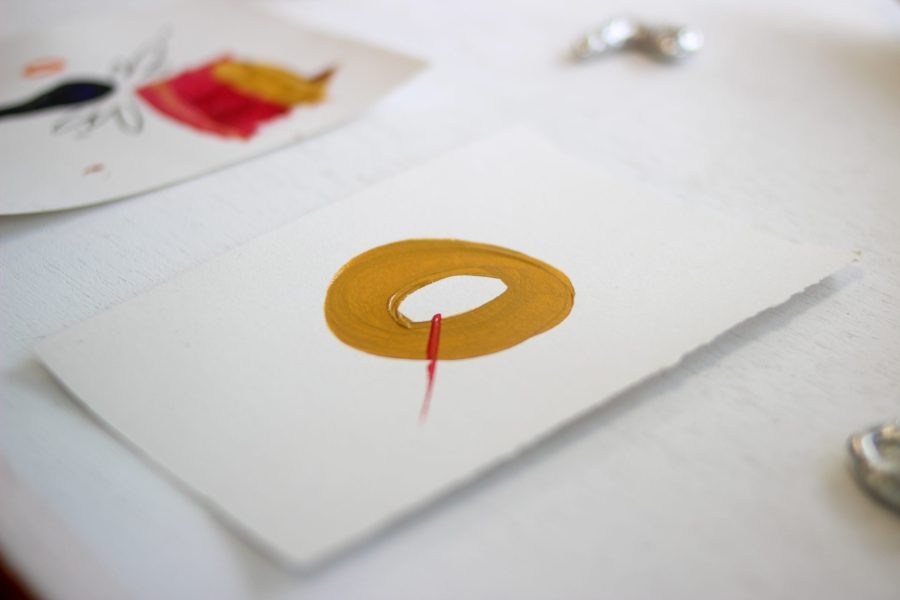 An artwork of a burnt yellow circle painted on a white piece of paper with a red dash through the middle.