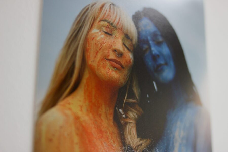 A close up of a photograph for exhibition Confine + Escape. A woman painted in orange with blonde hair and a fringe leaning and smiling with eyes closed close to a woman painted in blue with dark hair smiling with eyes closed.