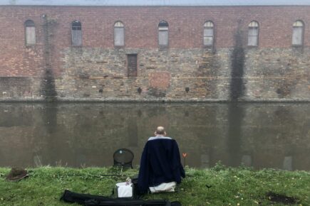 A fishermen sat at the Feeder Canal looking onto a brick building fishing with their back to the camera.