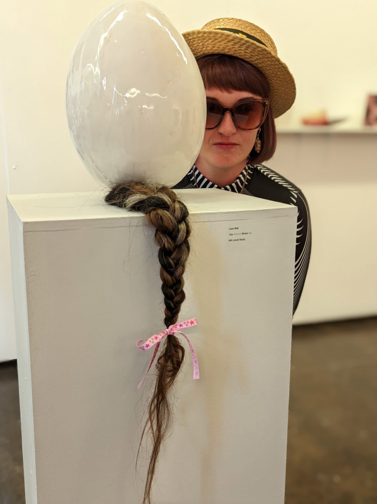 Artist Lara Bell in sunglasses and a hat, crouching behind a plinth with a balloon and hair on.