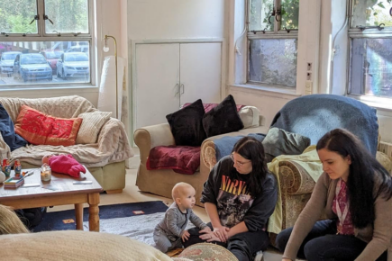 A young baby leaning on a woman's lap whilst another is bend down, they are surrounded by rugs and sofas.
