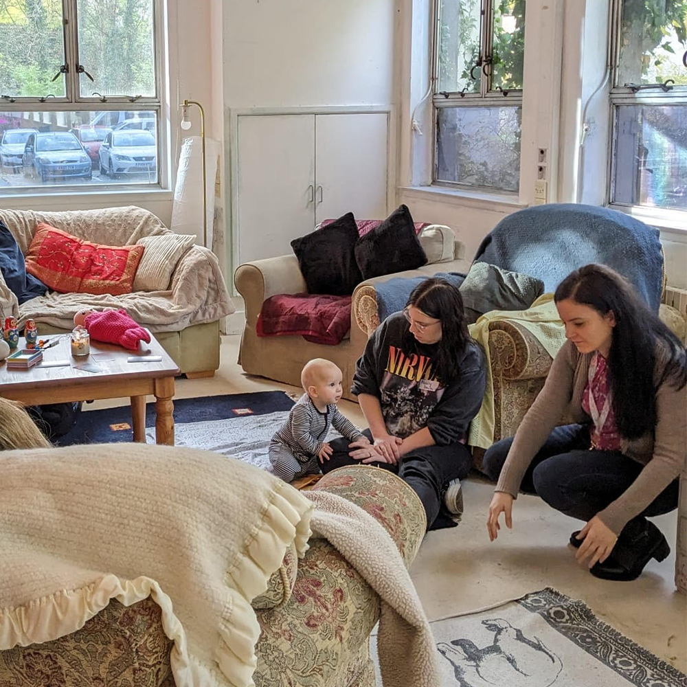 A young baby leaning on a woman's lap whilst another is bend down, they are surrounded by rugs and sofas.