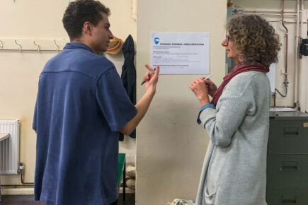 A young man in a blue top and brown hair talking to a woman and using hand gestures holding pens with a piece of paper on the wall reading ' Economy, business, jobs and education'.