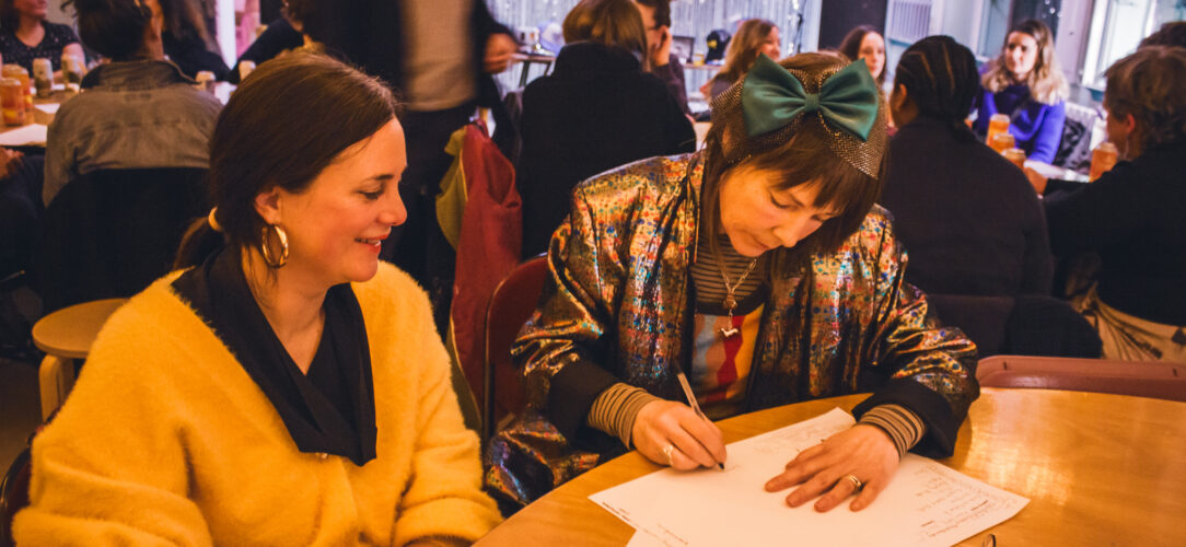Two women sat writing on pieces of paper smiling. The first one is in a yellow cardigan with gold hoop earrings, the second has a blue bow and shiny blazer on.