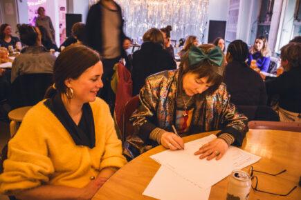 Two women sat writing on pieces of paper smiling. The first one is in a yellow cardigan with gold hoop earrings, the second has a blue bow and shiny blazer on.
