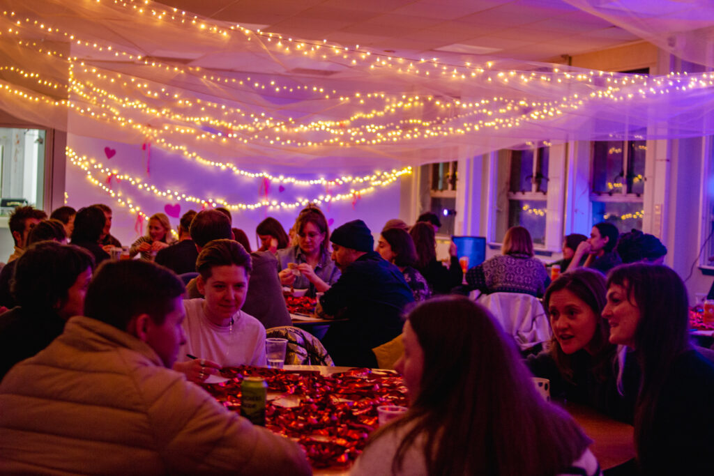 A room full of people sitting around tables chatting whilst a man in the far background speaks into a microphone. The room is filled with fairy lights and soft fabric.