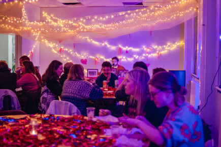 A room full of people sitting around tables chatting whilst a man in the far background speaks into a microphone. The room is filled with fairy lights and soft fabric.