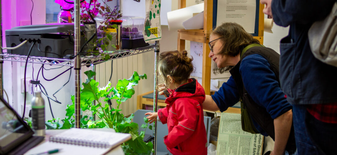 A toddler in a red jacket pointing at a plant growing in a metal stand with their mother leaning over and looking too.