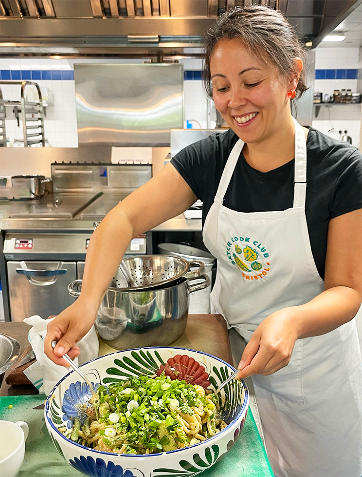 A woman with black hair, smiling in a black t-shirt and white apron mixing a colourful bowl of green and healthy food.