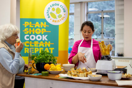 A woman with dark hair and a pink top on and apron, holding a blender with focaccia, lemons and oranges on the table in front of her. With an older women in blue with grey hair eating some focaccia. There's a standing banner behind them in blue and yellow that says 'Batch Cook Club, Bristol, Plan (crossed out), Shop (crossed out), Cook, Clean (crossed out), Repeat'