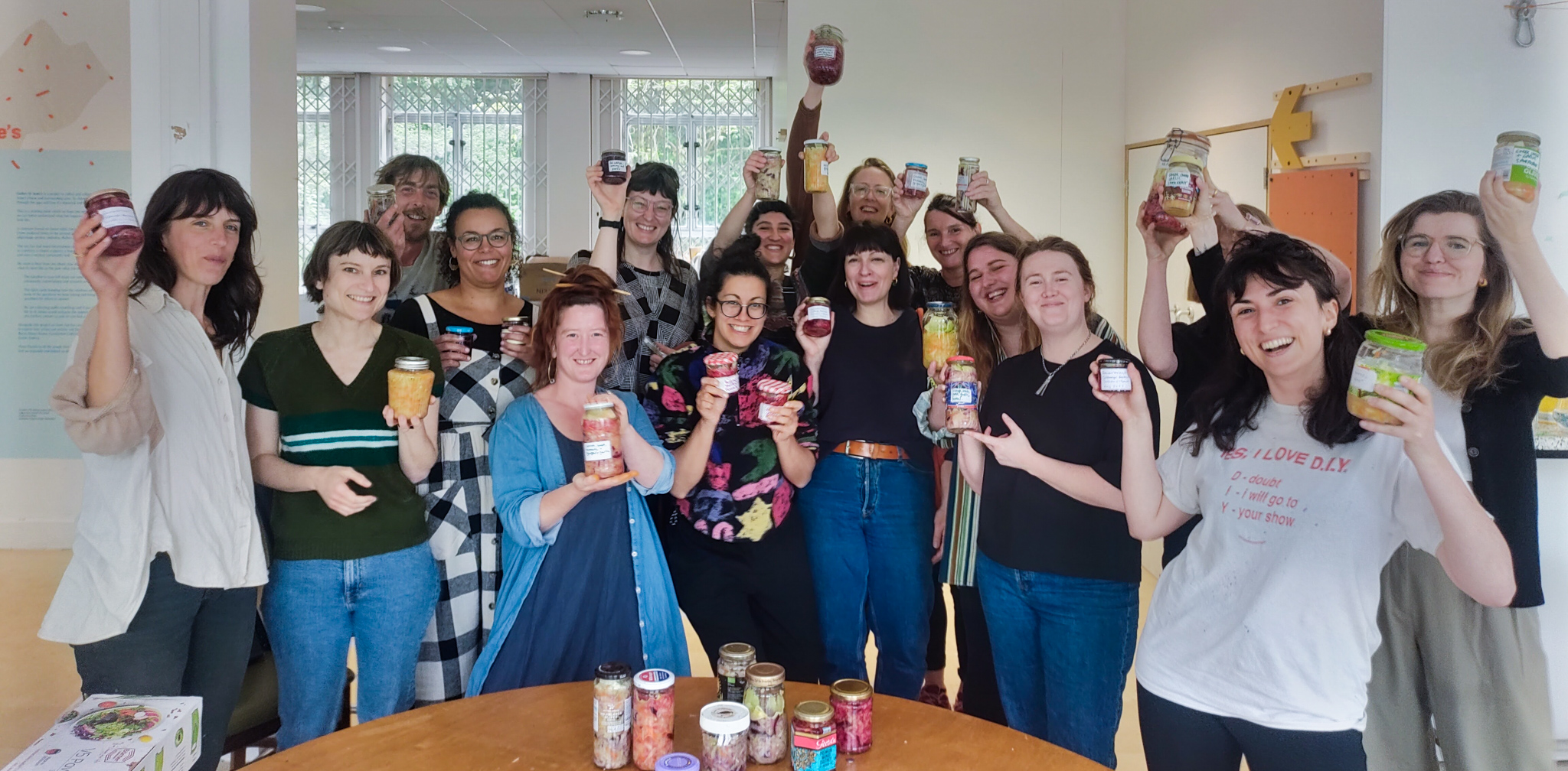 Around 12 people are smiling at the camera and raising their hands up holding jars that they have pickled at St Anne's House for Jar squad.