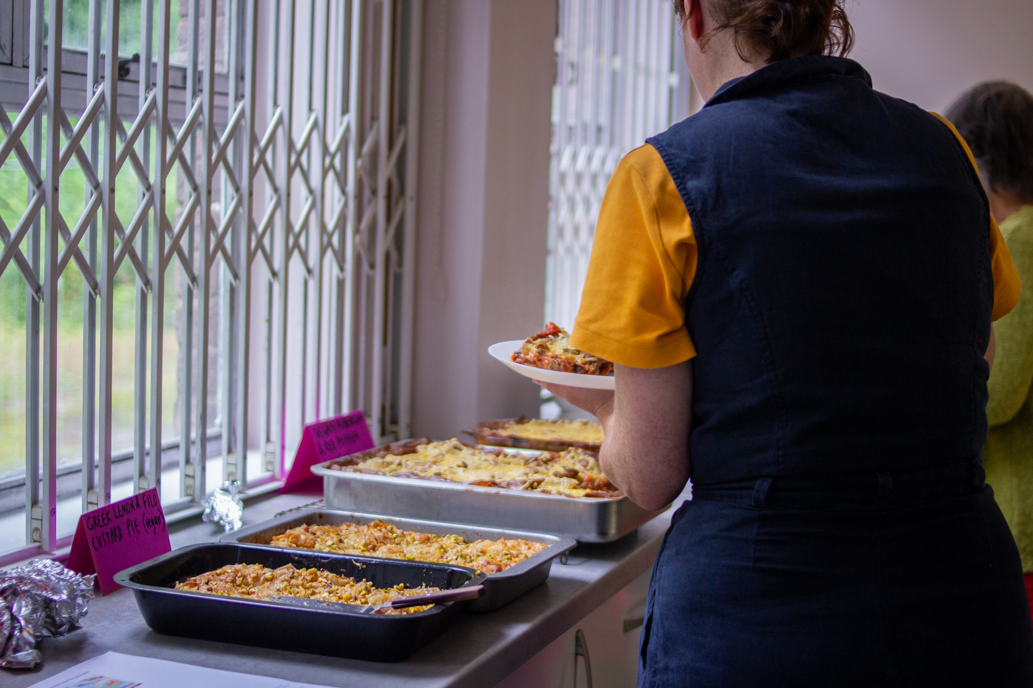 Woman with her back to us holds a plate of lasagne and stands in front of trays of food.