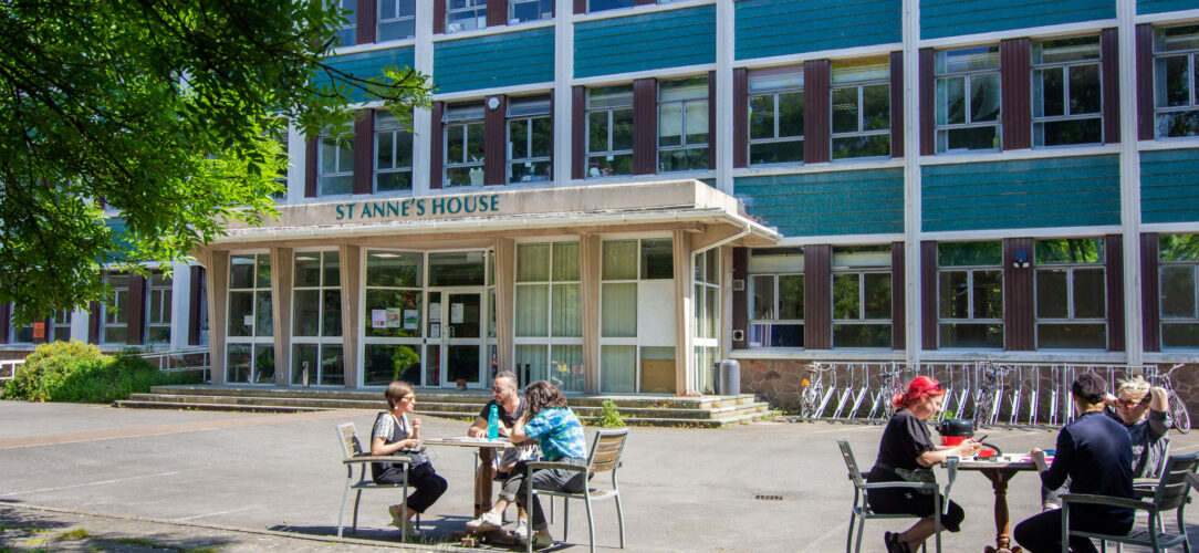 A large building with teal blue tiling and wooden panelling called St Anne's House, with two tables of different people sat outside chatting in the sun.