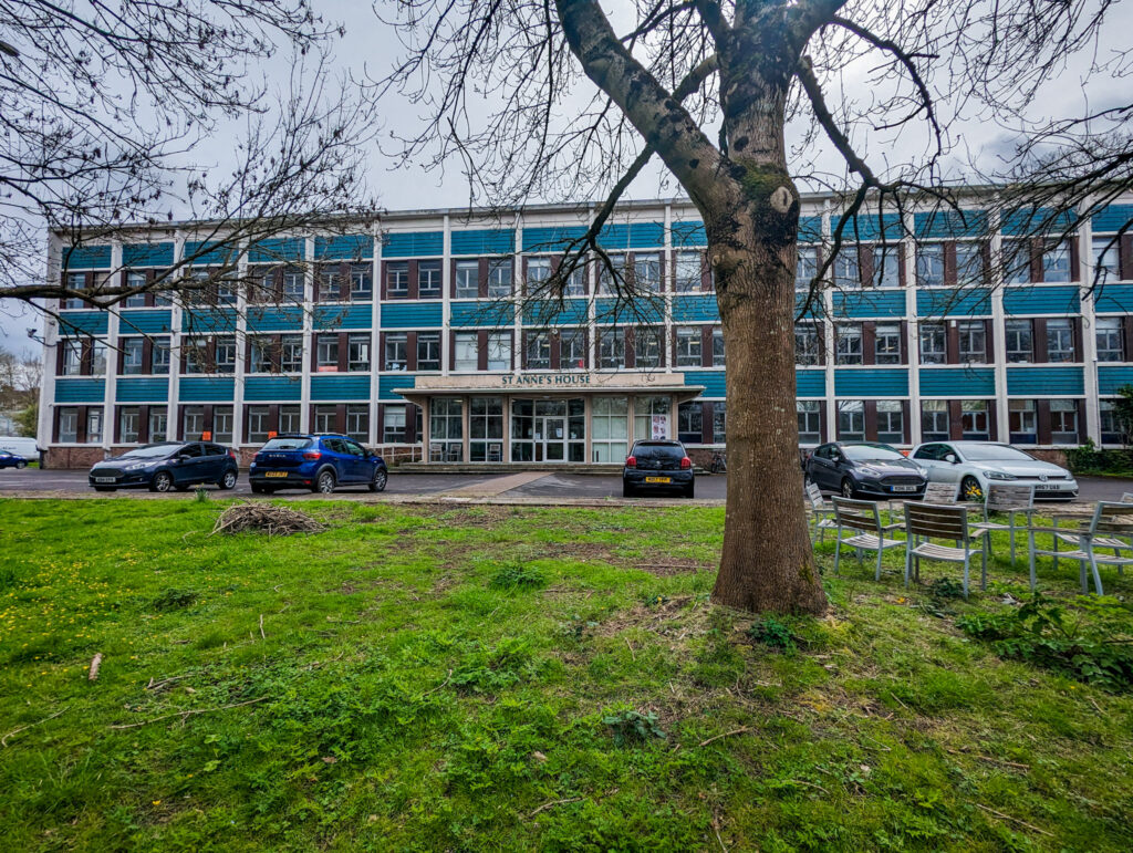 A patch of grass with a tree and behind a large building with lots of windows and teal tiling