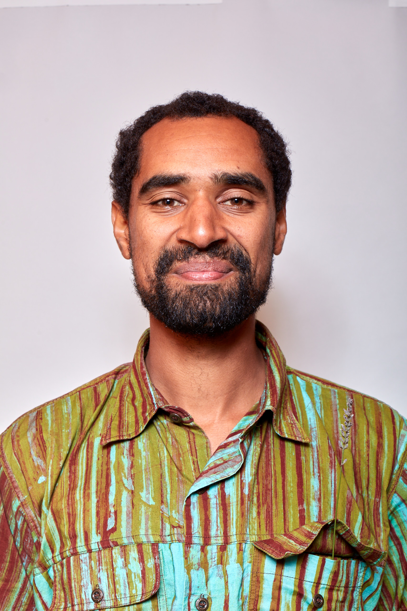 A head-and-shoulders portrait of a man standing against a plain light background. He has short curly hair, a trimmed beard and moustache, and is smiling gently at the camera. He is wearing a patterned button-up shirt with vertical stripes in green, brown and turquoise tones. The lighting is soft and even, giving the image a calm, natural feel.
