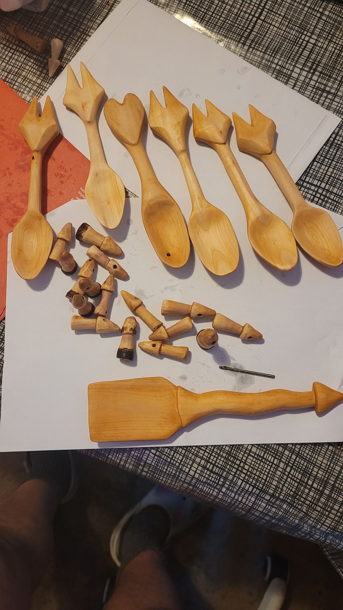 A set of hand-carved wooden utensils laid out on white paper on a patterned table. Seven small wooden spoons with decorative, crown-like carved handles are arranged in a row. Below them are several small carved wooden pegs or components, some with holes, scattered loosely. At the bottom is a larger flat wooden spatula with a tapered handle. The wood is light-coloured and unfinished, showing visible grain and hand-crafted details.
